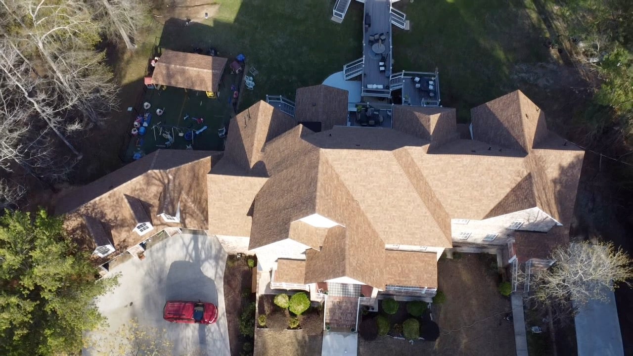 Aerial view of a large tan-roofed house, red car, decks, lawn, outdoor furnishings, and surrounding trees.